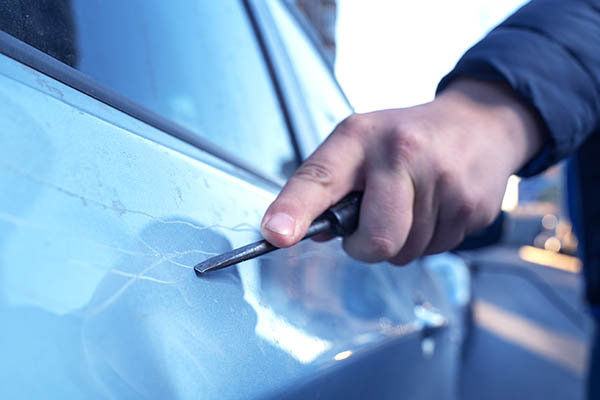 Hand with a screwdriver or a large nail scratching the paint surface on the vehicle door. A very stereotypical and perhaps a bit of ridiculous act of revenge.